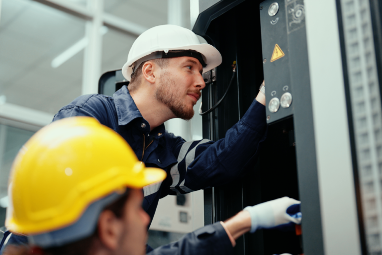 Two technicians in hard hats inspecting an industrial machine control panel in a manufacturing facility