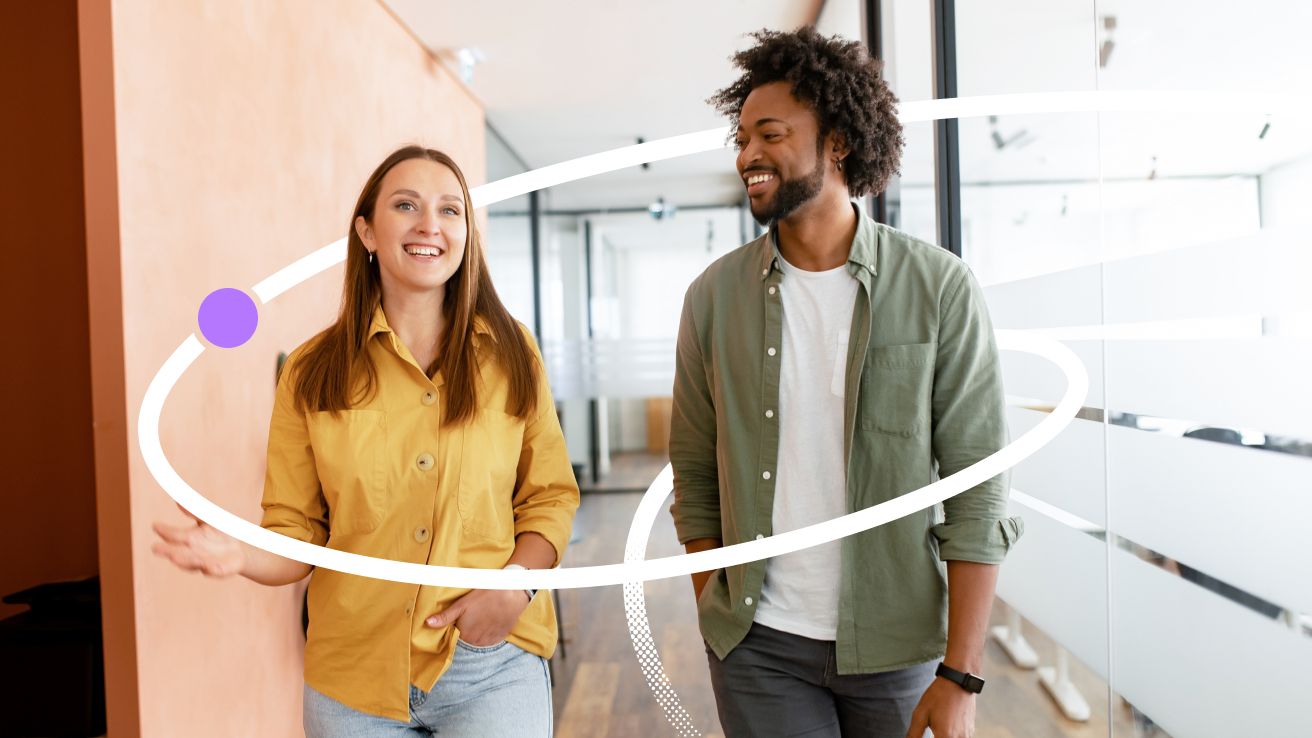 Two colleagues talking and smiling as they walk together through a bright, modern office hallway.